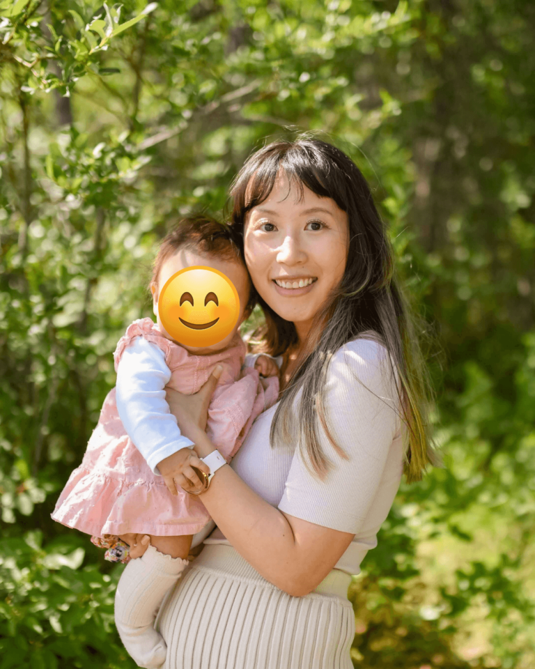 Emi Linds from Canada smiling while holding her young daughter outdoors, surrounded by sunlight and green foliage.