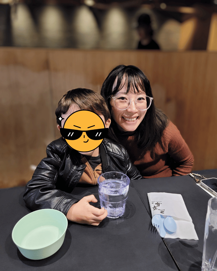 Emi Linds sitting next to her son at a restaurant table, smiling warmly with glasses and a casual outfit