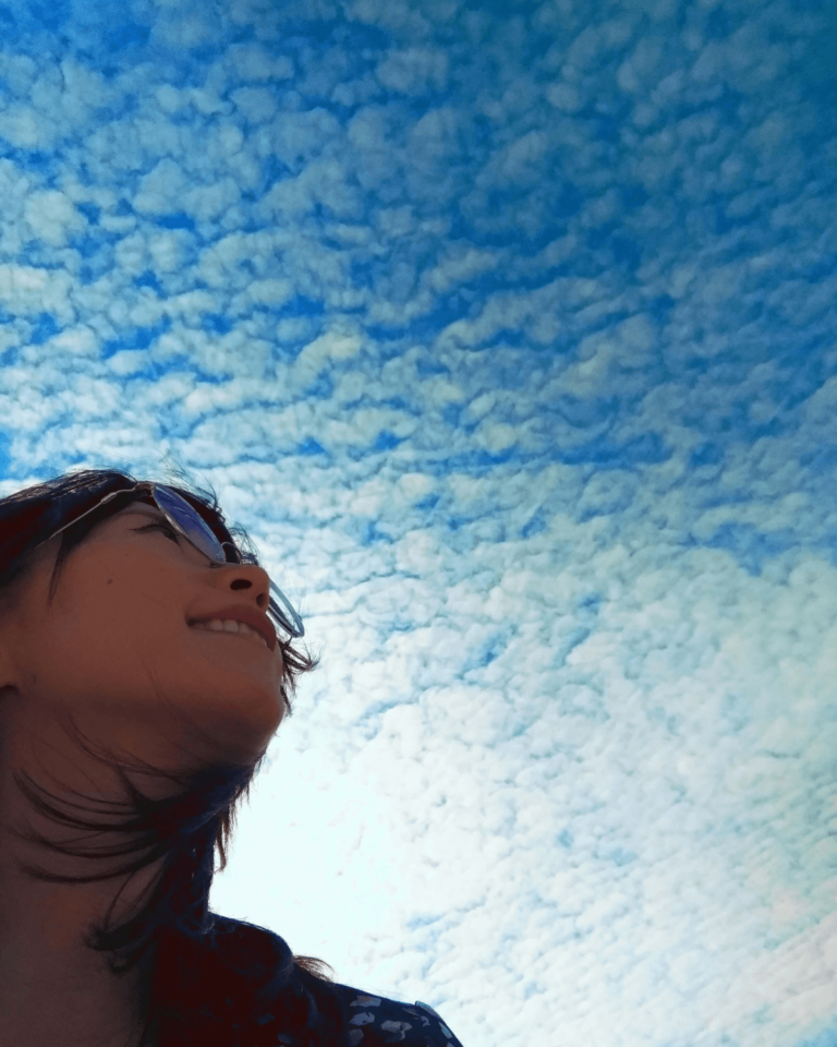 Emi Linds, partially in frame, looks upward under a rippling sky of cloud and light, wearing sunglasses and a subtle smile.